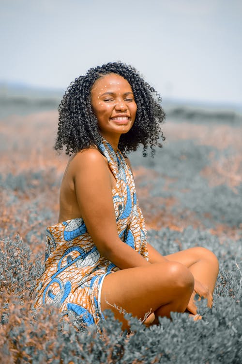 Side view of happy African American female with Afro hairstyle sitting in meadow and smiling happily at camera