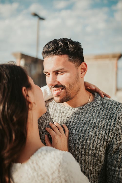 Young couple in casual clothes standing on street together under blue cloudy sky in sunny summer day and gently embracing while looking at each other