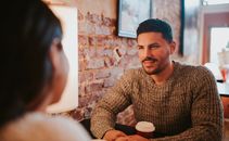 Young couple in casual clothes sitting at table with cup of drink on date in cafeteria near brick wall and talking