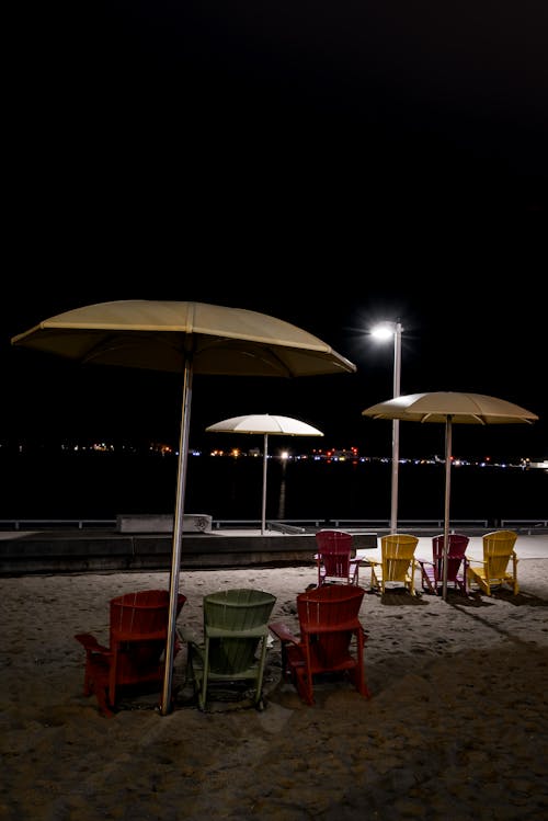 Sandy coast with streetlamp and colorful plastic deckchairs near umbrellas placed on embankment against glowing lights of city at night time