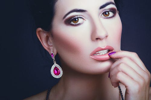 Charming female with bright makeup in stylish accessories touching lips and looking at camera against dark background