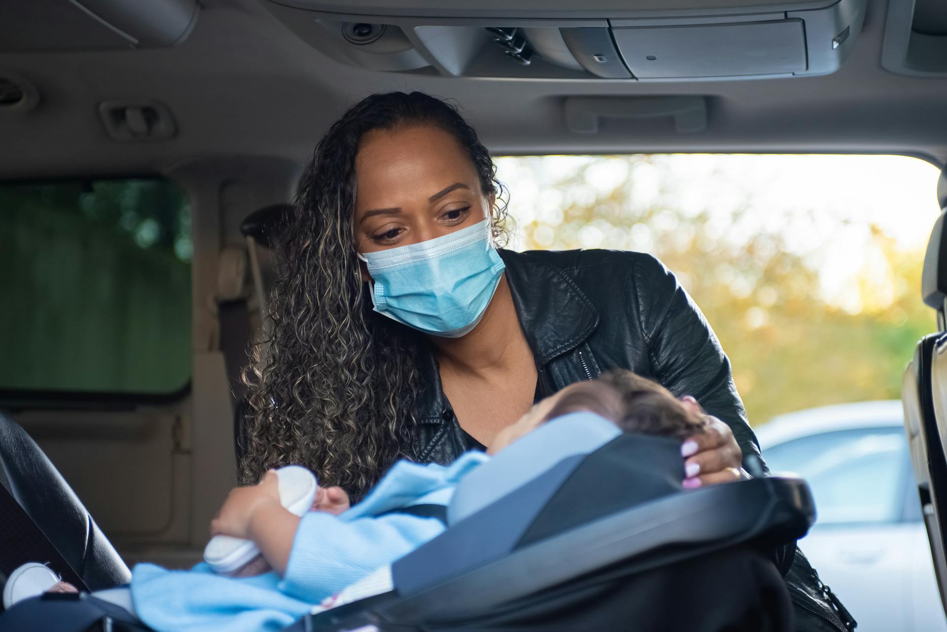 A caring mother wearing a face mask buckles her baby into a car seat, promoting safety and love.