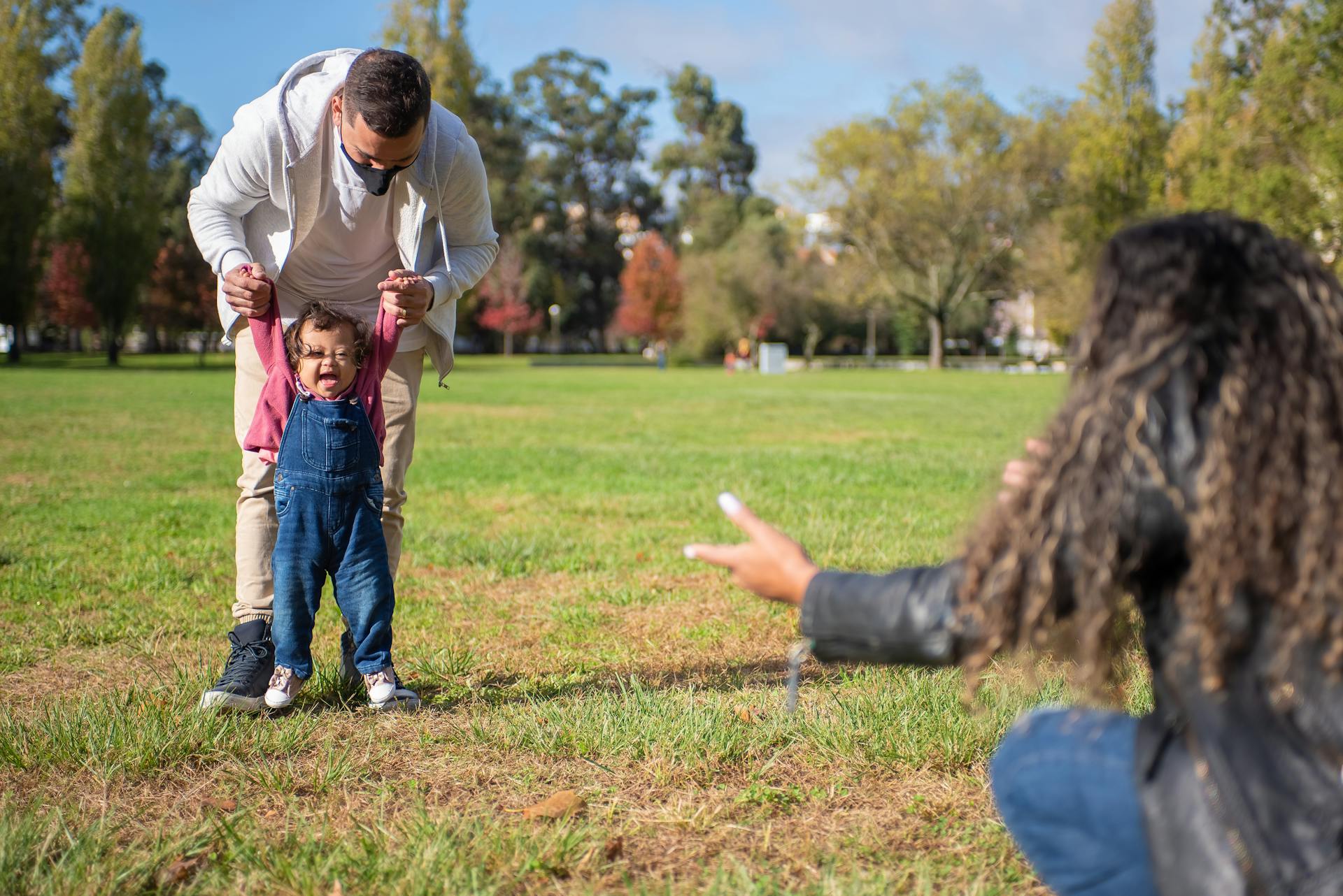 Father supporting his baby taking first steps while mother reaches out in a sunny park.