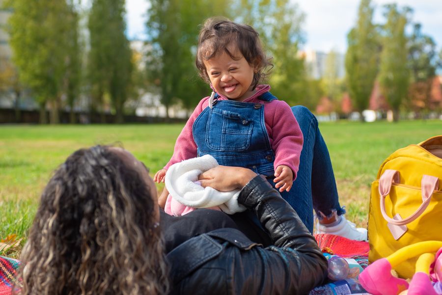 Mãe sorrindo e passando tempo com a filha com síndrome de Down durante um piquenique no parque, imagem ilustrativa de acolhimento e parentalidade atípica