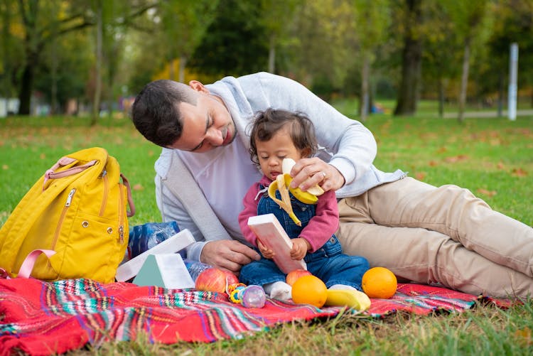 
A Man Feeding His Child With A Banana While On A Picnic Blanket