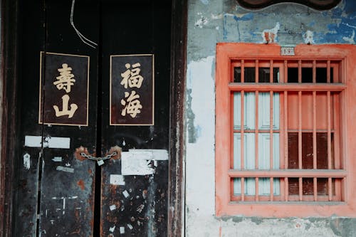 Old shabby door with written hieroglyphs and red window with metal grid on wall of aged building on street in city