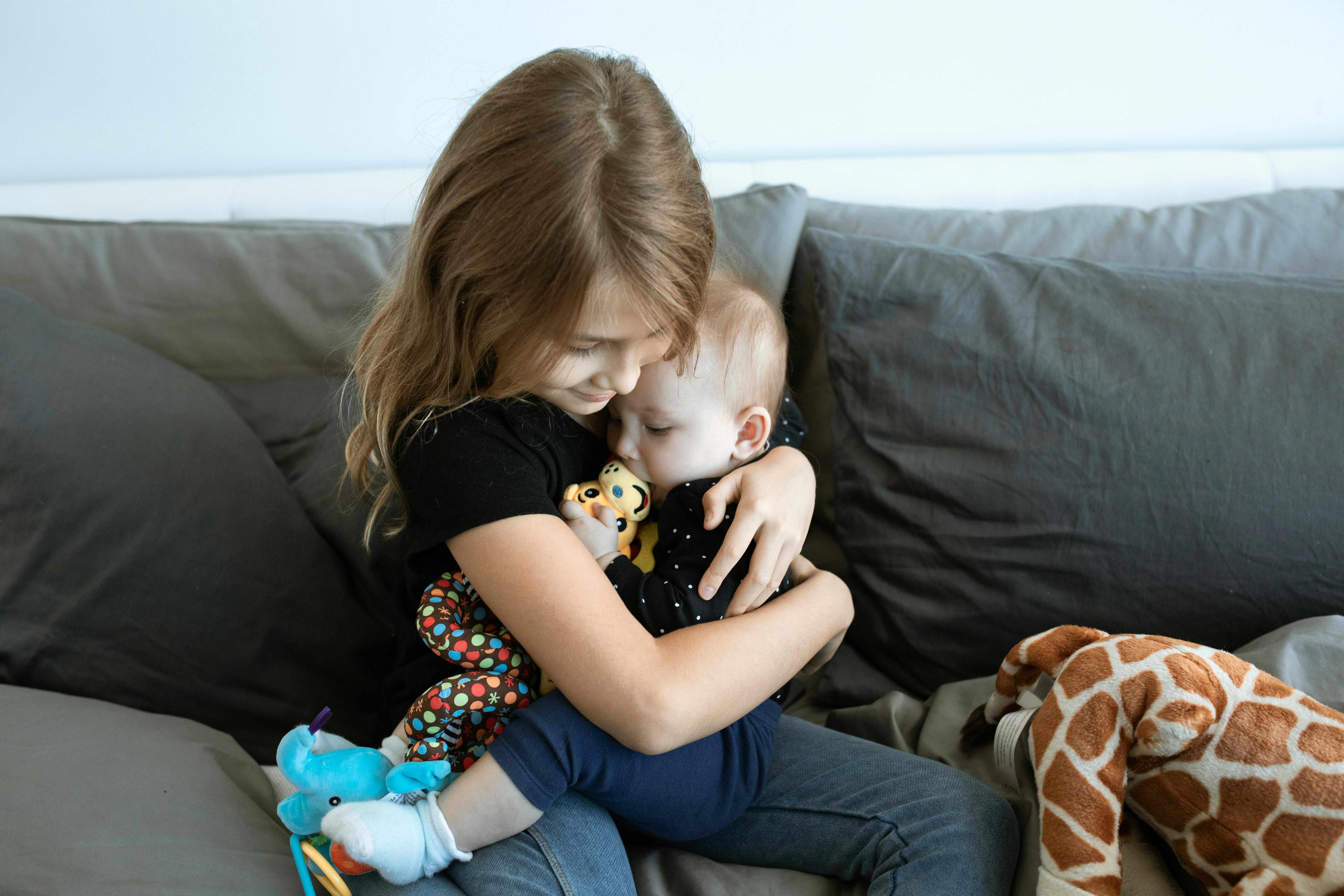 girl in black t shirt and blue denim jeans sitting on grey sofa holding her baby brother