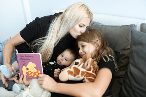 A Mother Kissing the Hair of Her Daughter Reading a Book for Her Baby Sister