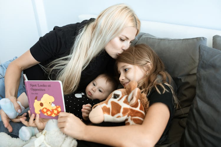 A Mother Kissing The Hair Of Her Daughter Reading A Book For Her Baby Sister