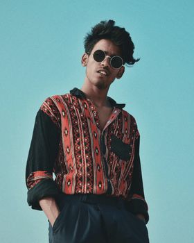 A stylish man in retro attire poses confidently outdoors with sunglasses.