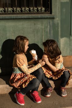Two young girls sharing ice cream cones while sitting outdoors in the summer sun.