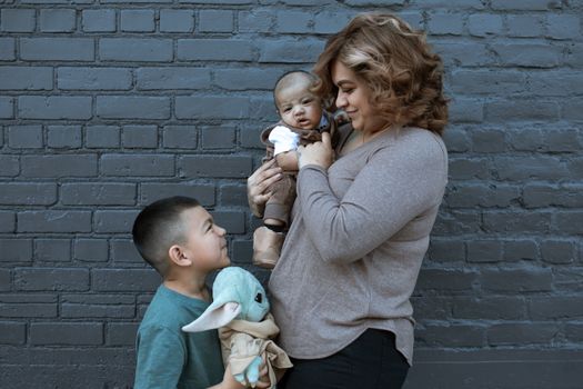 A joyful family moment with a mother holding a baby and a young boy holding a plush toy against a gray brick wall.