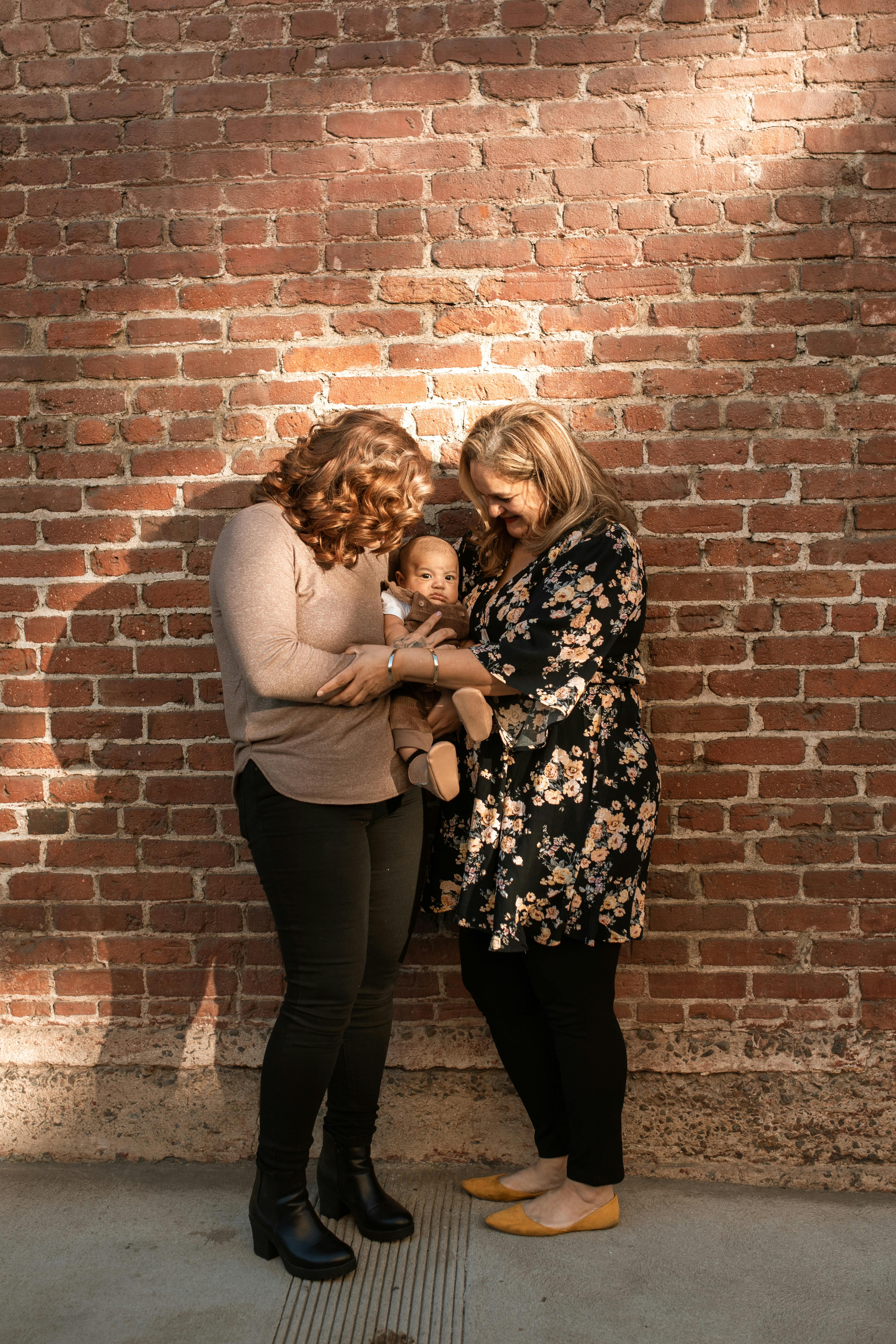 two women standing beside brick wall holding a baby
