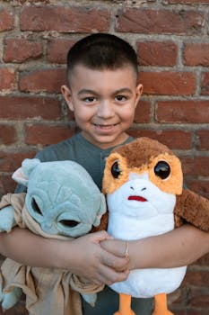 Young boy happily holding Baby Yoda and Porg plushies against a brick wall.
