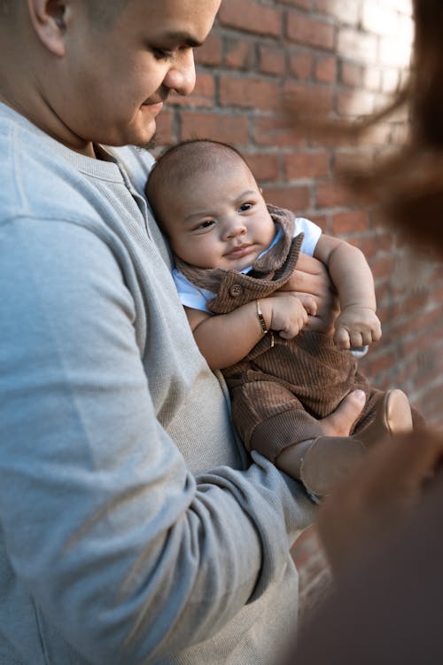 Free A Man in Gray Shirt Carrying a Baby Boy Wearing Brown Jumper Stock Photo