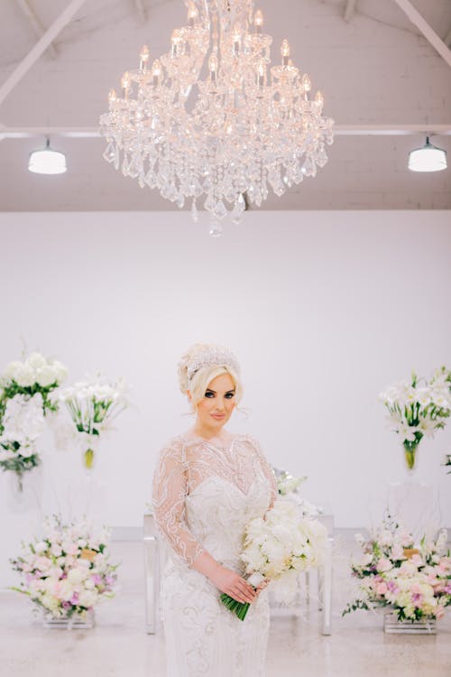 Free A Beautiful Bride Standing Under the Chandelier while Holding a Bouquet of Flowers Stock Photo