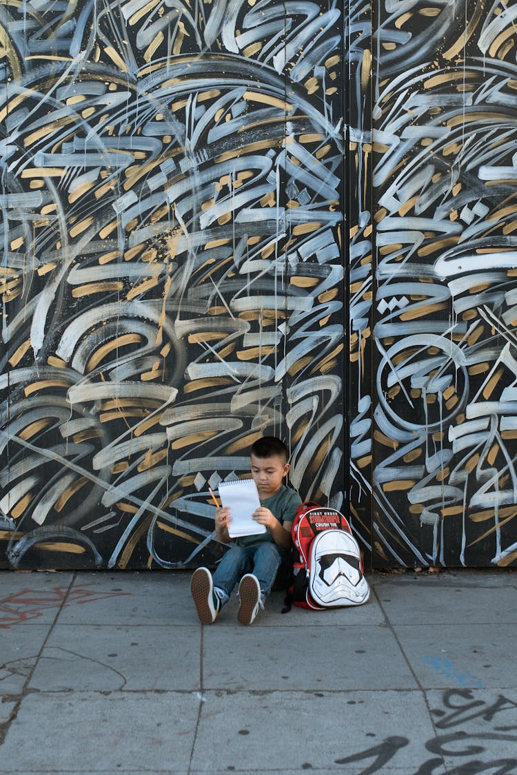 A Young Boy Sitting On The Street While Holding His Notebook