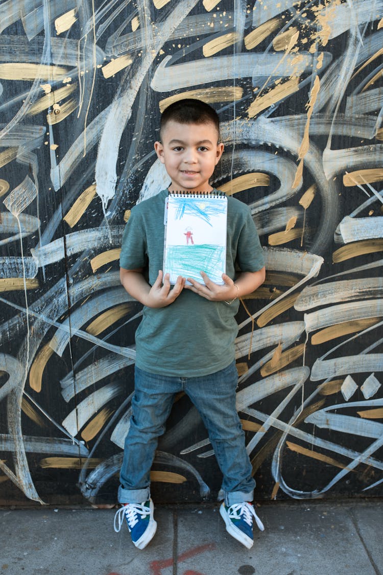 A Young Boy Standing On The Street While Holding His Notebook With Drawing