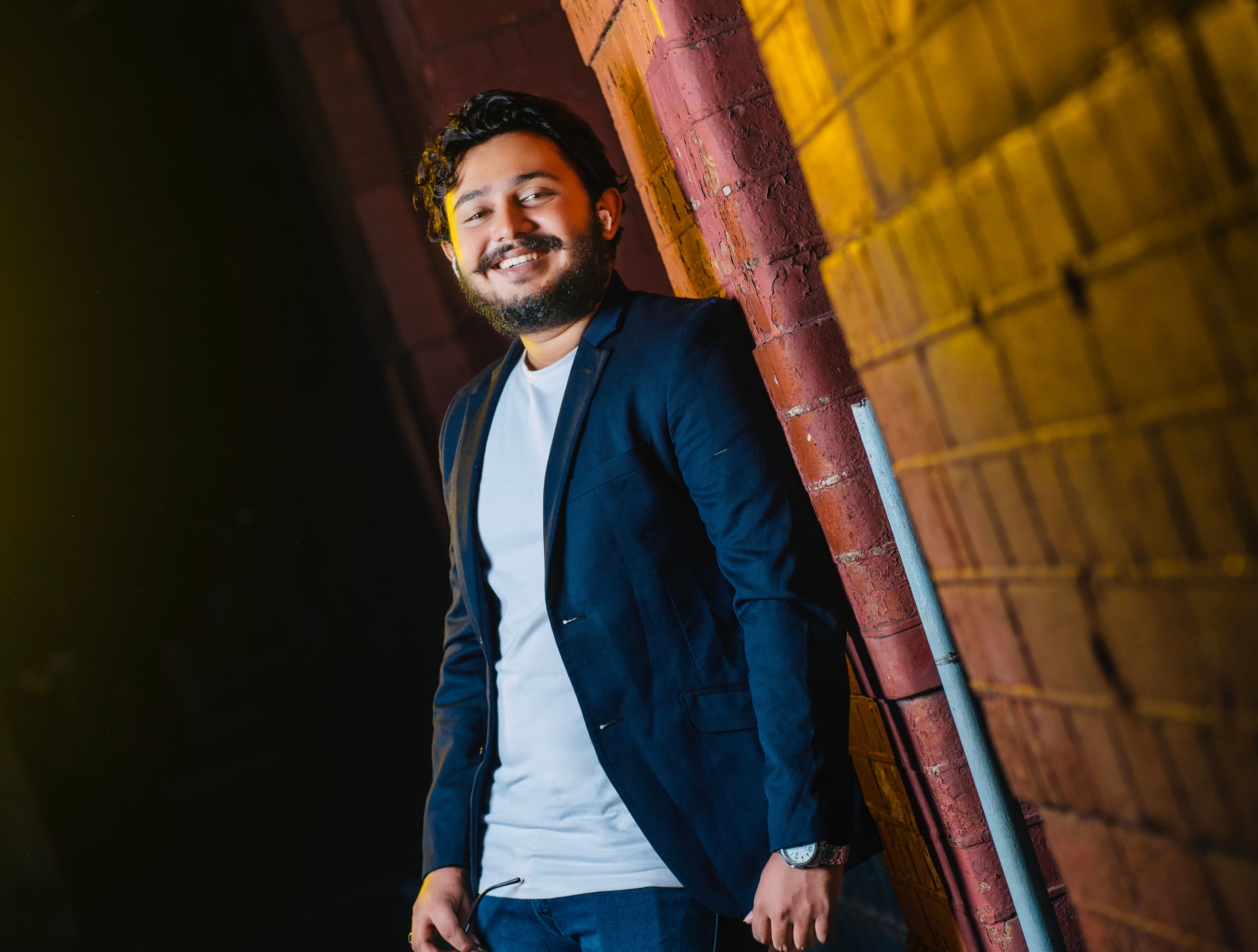 A bearded man in a blazer smiles as he leans against a brick wall. Captured in Sri Lanka.