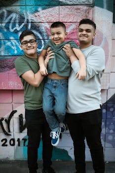 Two older boys lift younger brother in front of colorful urban mural, smiling.