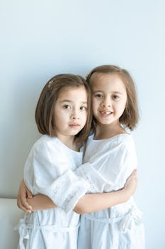 Adorable twin sisters embracing, wearing white dresses in a minimalist indoor setting.