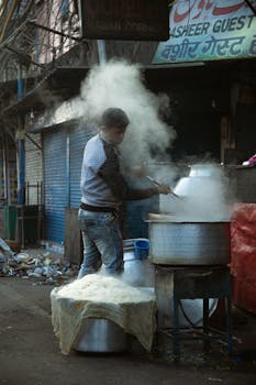 A street vendor stirring a steaming pot in an urban alleyway, wearing denim and long sleeves.