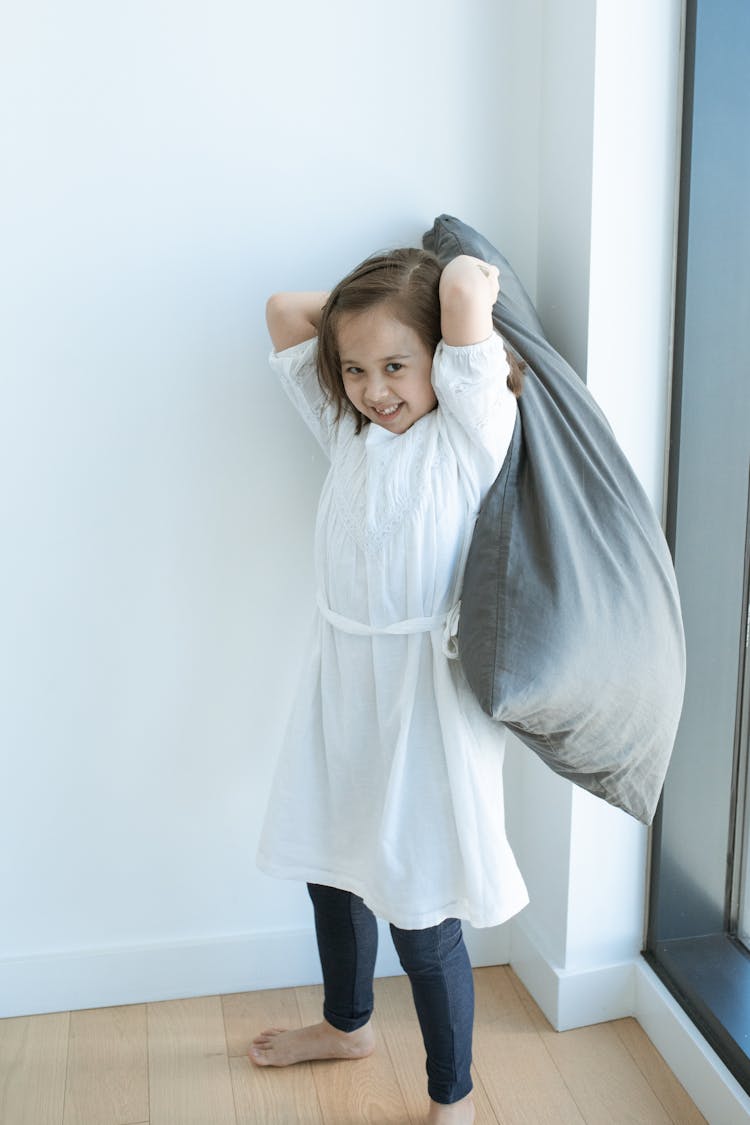 A Cute Girl In White Dress And Leggings Holding A Gray Pillow