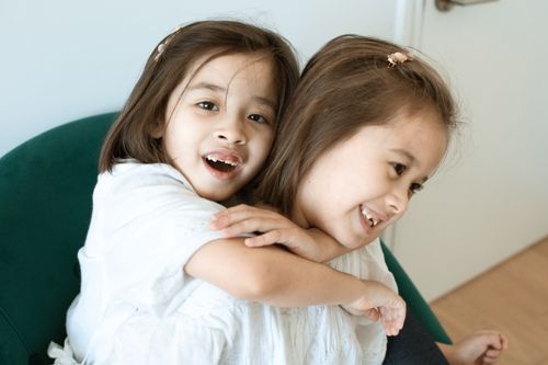 Free Two smiling young girls hugging warmly on a chair inside a cozy room. Stock Photo