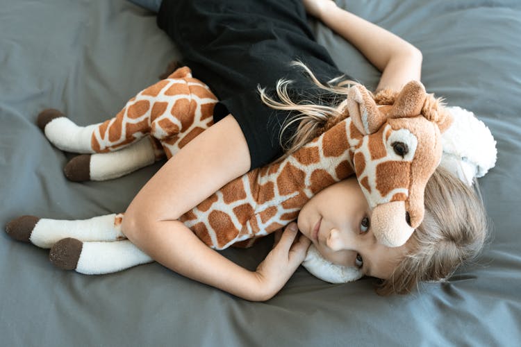 Overhead Shot Of A Kid Lying Down On A Bed With Her Giraffe Stuffed Toy