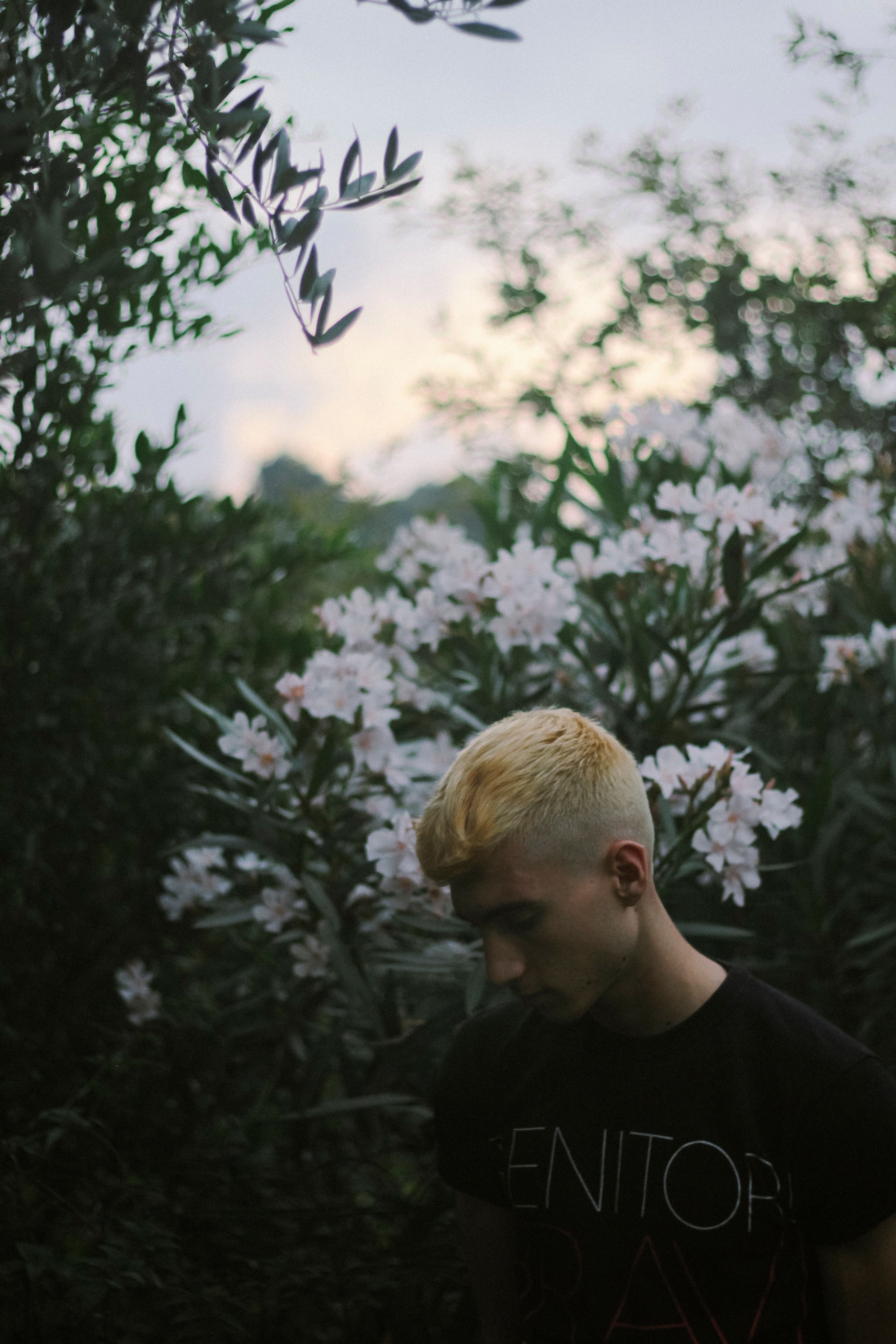 A young man with blonde hair stands amidst lush flowering shrubs, looking down pensively.