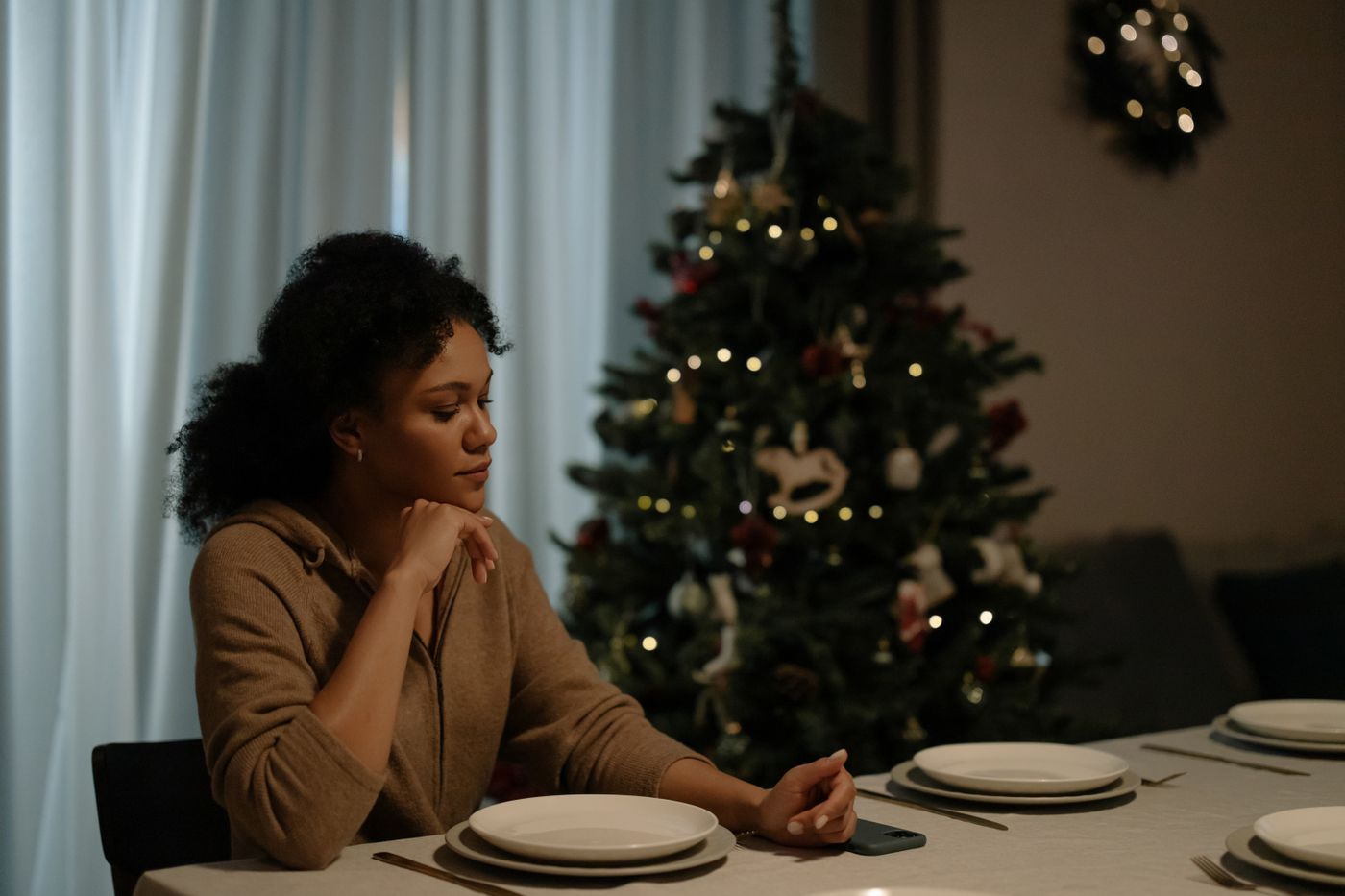 A woman sitting alone at a set dinner table, a lit Christmas tree behind her.