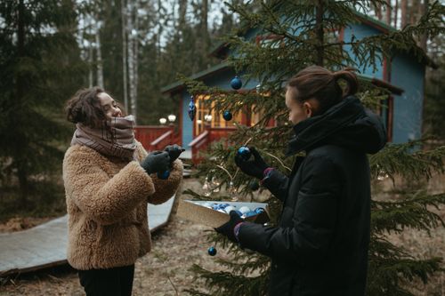 Free Two friends enjoy decorating a Christmas tree outdoors near a cozy cabin. Stock Photo