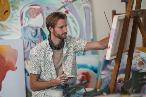 Free Bearded male artist painting on canvas in a vibrant studio setting, surrounded by artwork. Stock Photo