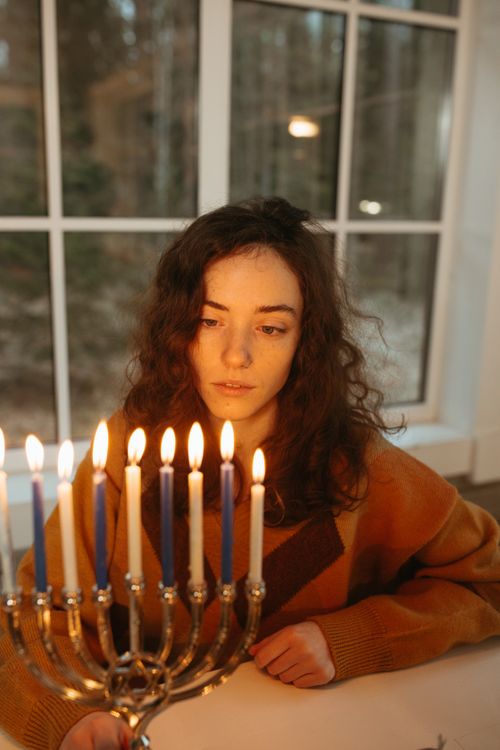 Free A woman in a sweater gazes at a lit menorah near a window, creating a warm, reflective atmosphere. Stock Photo