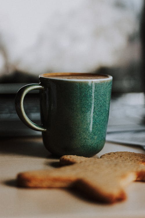 Green ceramic cup with fragrant fresh coffee and tasty crispy gingerbread cookie placed on table at home