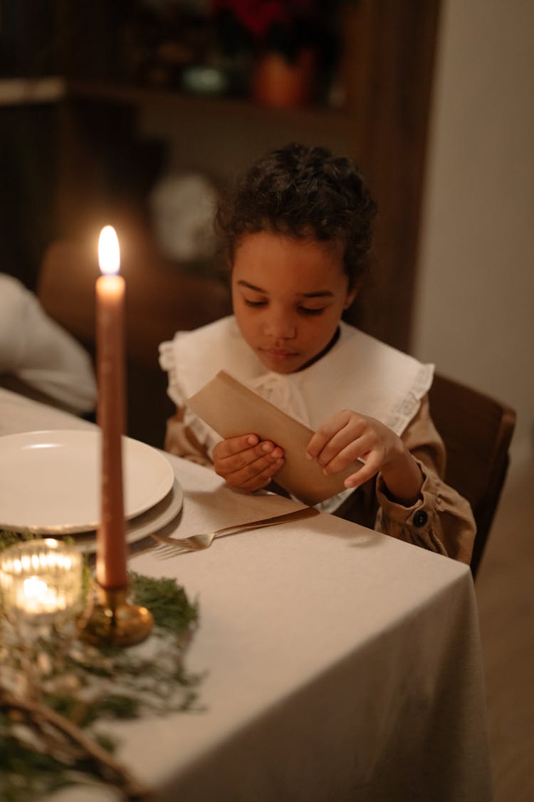 A Kid Sitting At A Dining Table Opening An Envelope