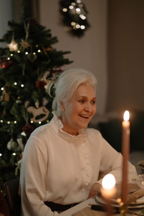 Free A joyful senior woman celebrating Christmas by a candlelit table with a decorated Xmas tree. Stock Photo