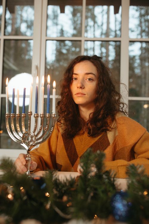 Free A woman in a cozy sweater celebrates Hanukkah indoors with a lit menorah, creating a warm festive atmosphere. Stock Photo
