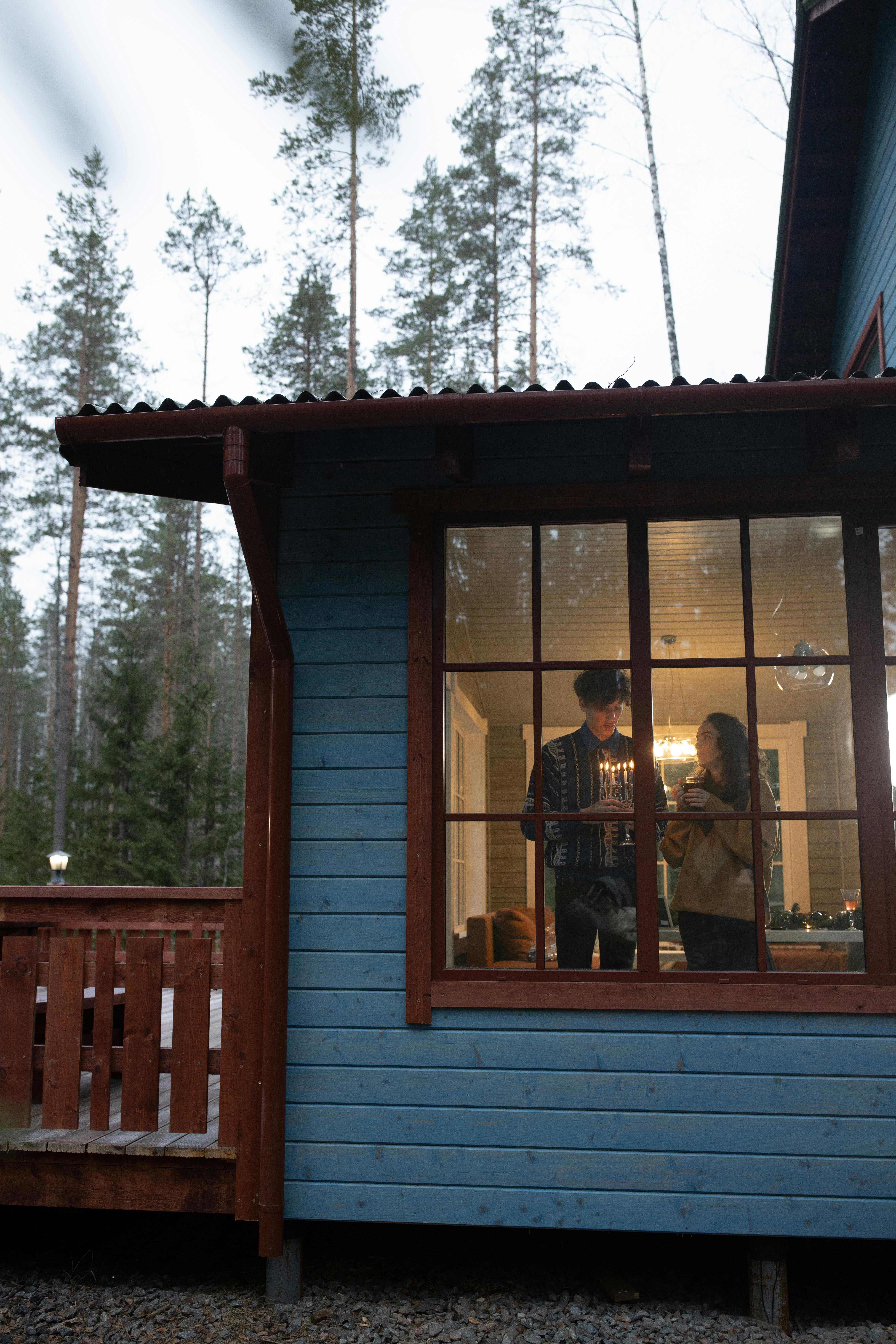 A couple enjoys a festive Hanukkah evening indoors, seen through a window of a rustic cabin.