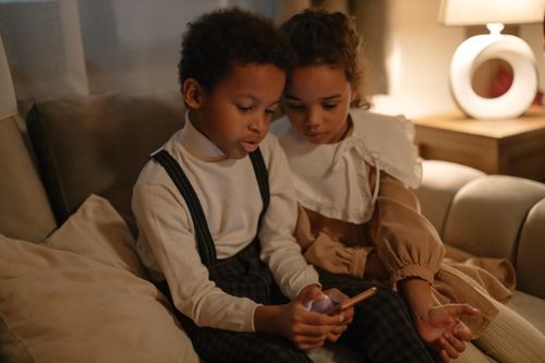 Free Two children sitting on a sofa, engaged with a smartphone during a cozy evening indoors. Stock Photo