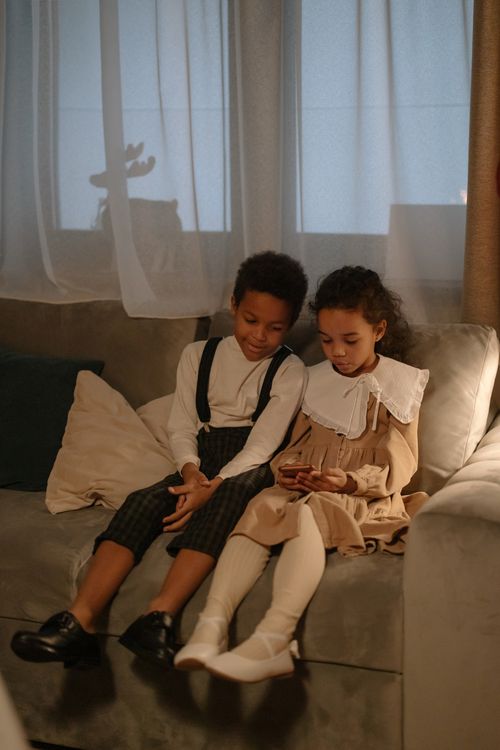 Free Young siblings sitting on a couch, sharing a smartphone indoors, enjoying technology and bonding. Stock Photo