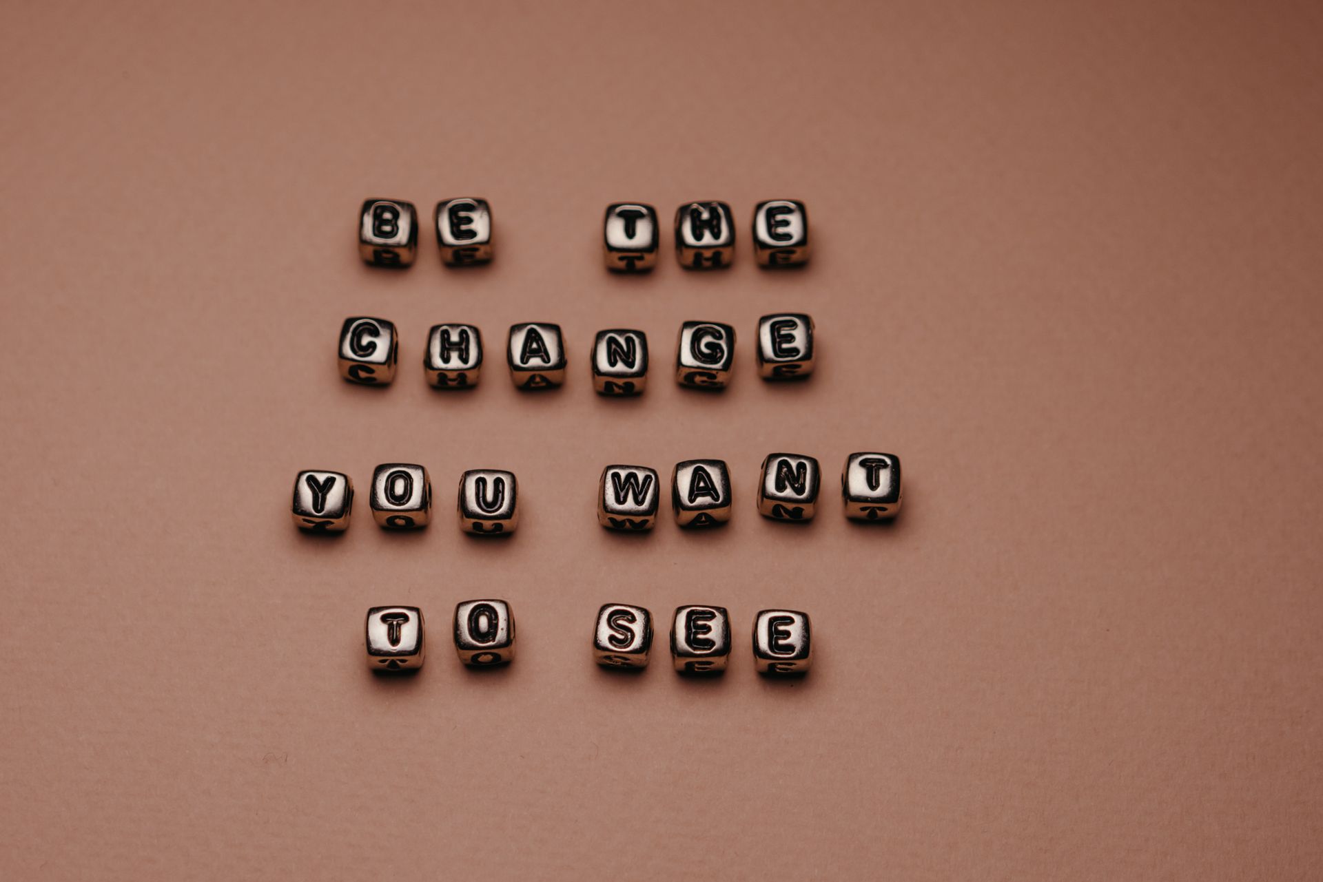 Inspirational message 'Be the change you want to see' displayed with alphabet beads on pink surface.