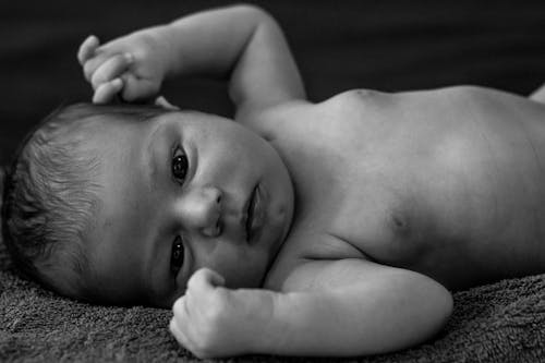 Black and white of cute naked newborn baby lying on soft blanket with raised arms and looking at camera