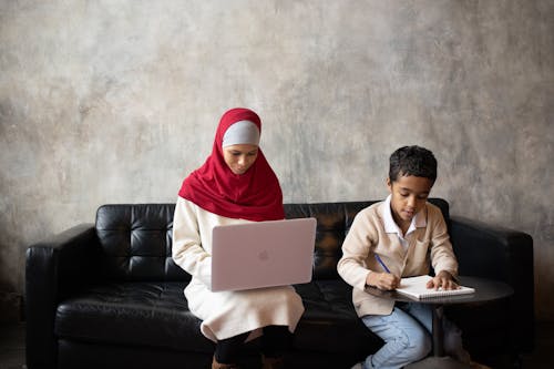 Focused Arabian son taking notes in copybook while mother working on laptop