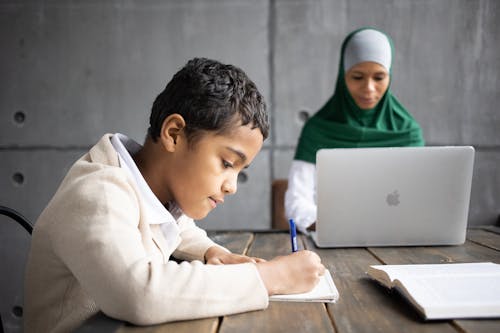 Serious Arabian boy writing homework while sitting at table with mother wearing traditional hijab working remotely on portable laptop