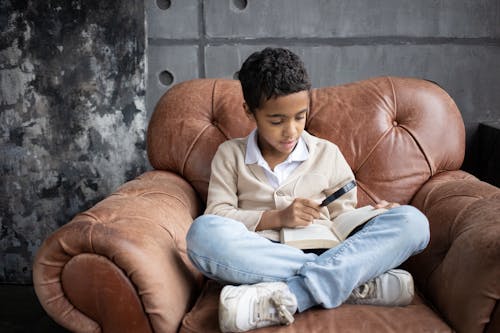 Full body of curious Arabian schoolboy sitting on comfortable armchair with crossed legs and reading book with magnifier at home