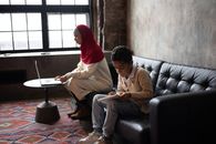 Side view of concentrated son preparing project for school while sitting on soft couch together with Arabian mother surfing laptop