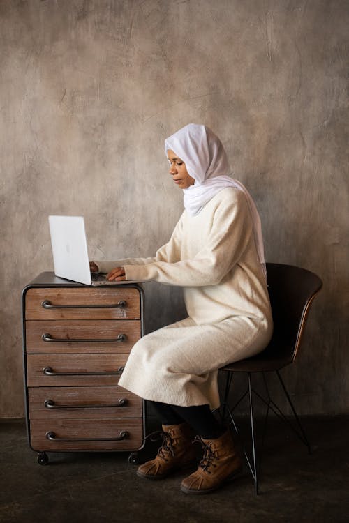 Side view of ethnic female in hijab typing on netbook while working on timber cabinet