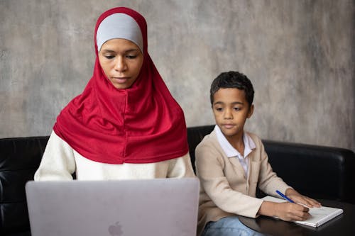 Muslim pensive ethnic mother typing on netbook while son with pen taking notes in notebook