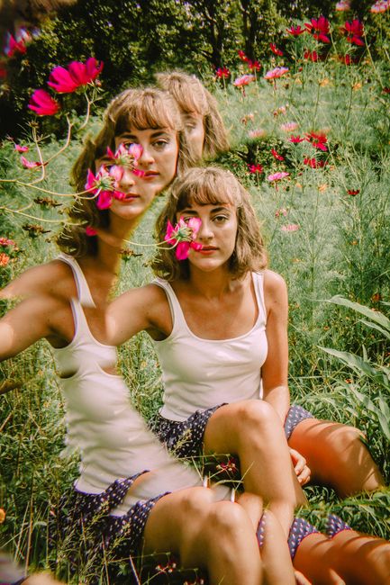 A woman in a white tank top reflects in a mirror among red flowers.